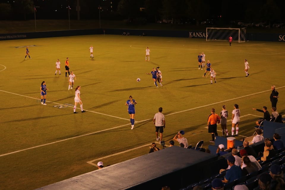 A nighttime soccer match in progress at Rock Chalk Park with KU's Women Soccer Team in blue and the opposition in white.